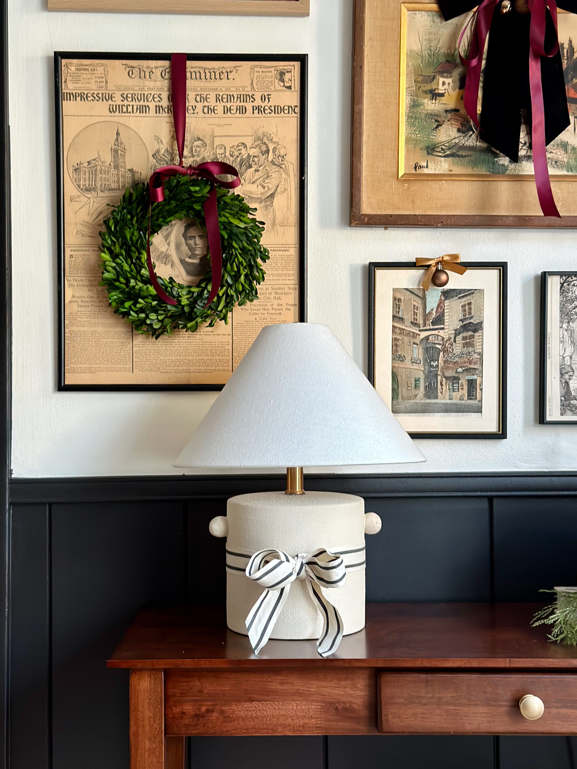 easy and budget friendly ways to decorate for christmas, decorating with bows, bow on lamp, decorating art for christmas, ribbon on art, wreath on art, christmas decor ideas, black wainscoting, mopboard black by benjamin moore, swiss coffee paint color, white paint, thrifted art, decorating gallery wall